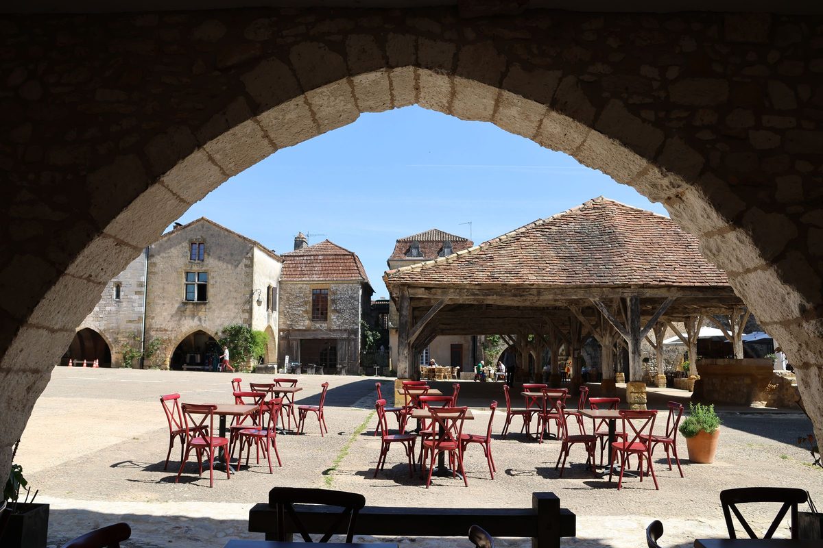 A sunlit village square seen through a stone archway, with empty red café chairs and a timbered market hall beyond