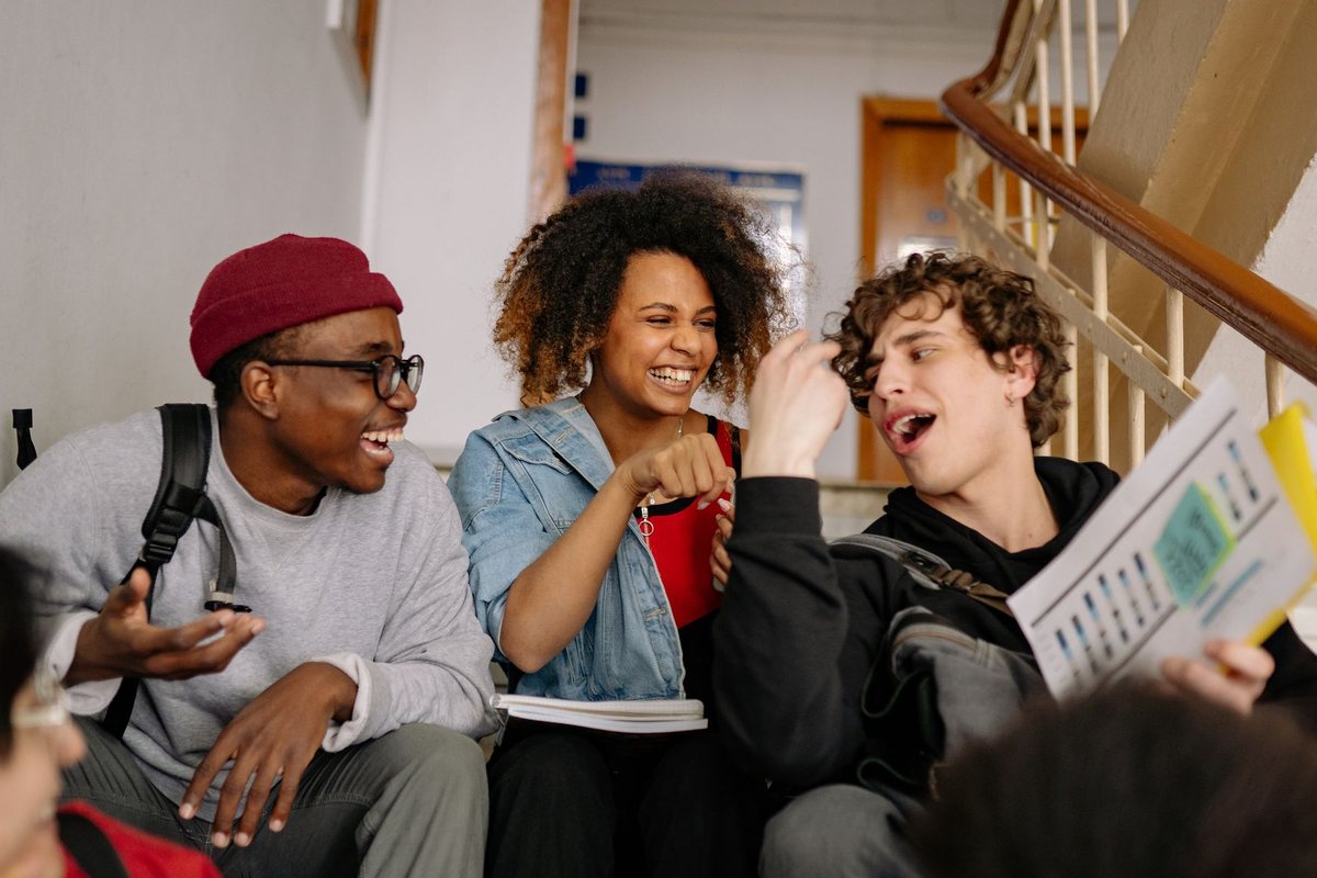 Three students laughing together on a school staircase, with classmates beside them