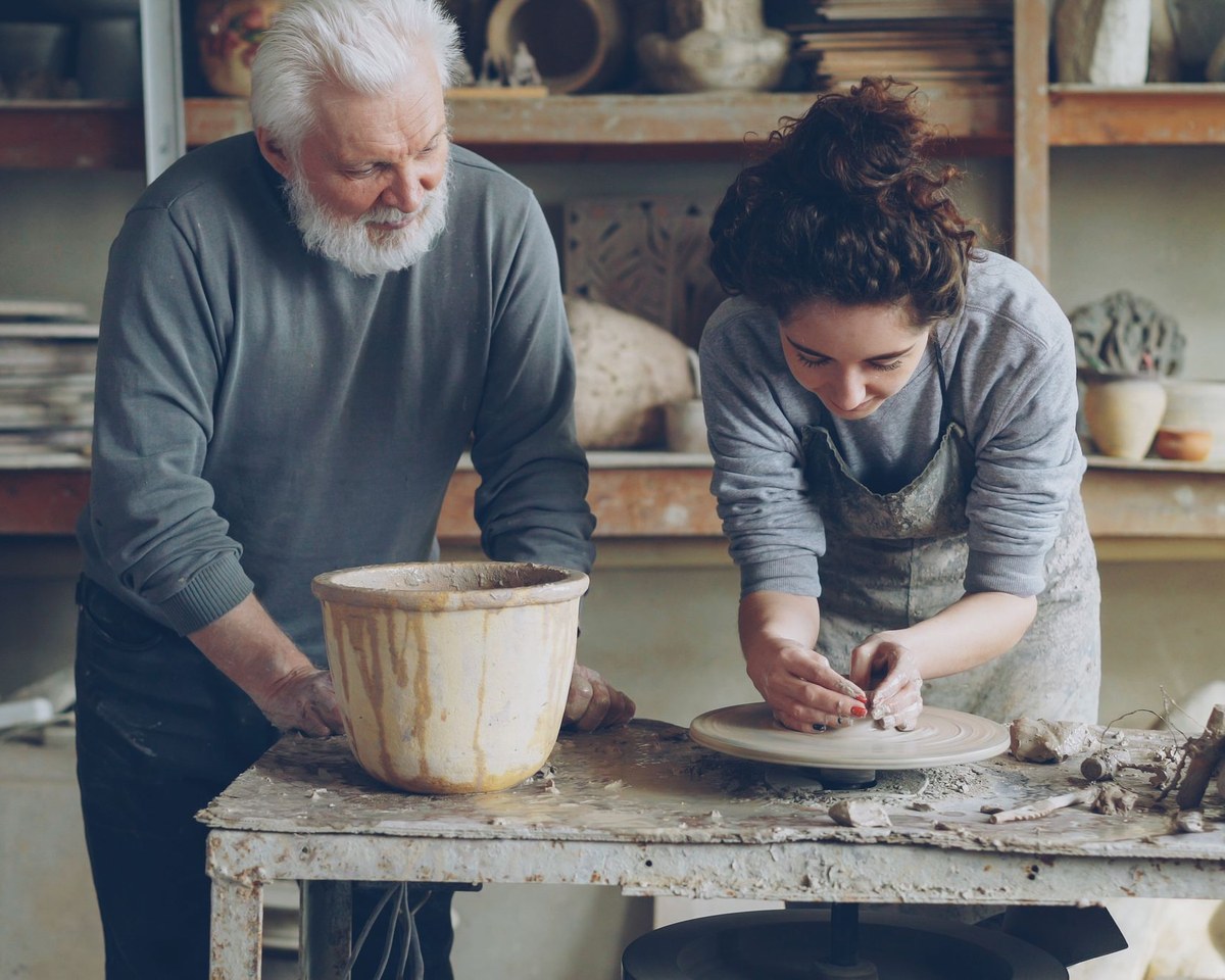 A grey-bearded older craftsman and a younger woman potter working together at a clay-covered table in a pottery studio, a large pot and a potter's wheel in front of them